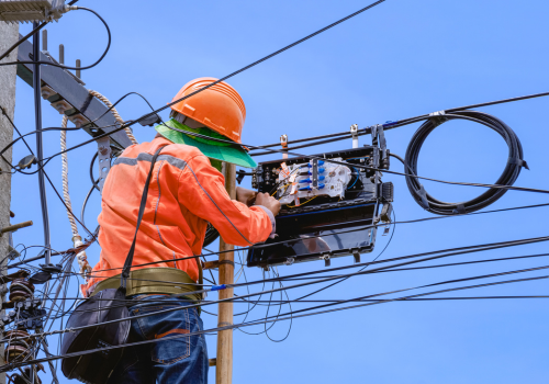 Technician on ladder is repairing fiber optic cable in internet splitter box on electric pole against blue sky background