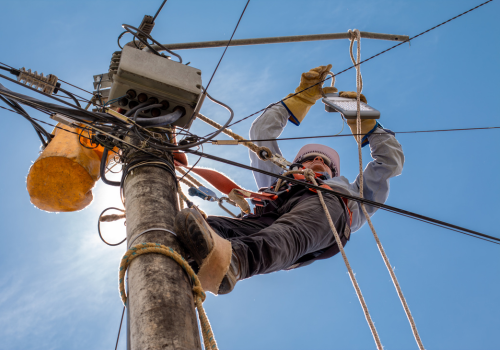 An electric utility lineman in a truck bucket is using a bolt cutter to cut a jumper wire connection on a high voltage power line. The worker is wearing insulated gloves and sleeves. The orange tubes are "guts" which are insulation to wrap around a power line for protection.